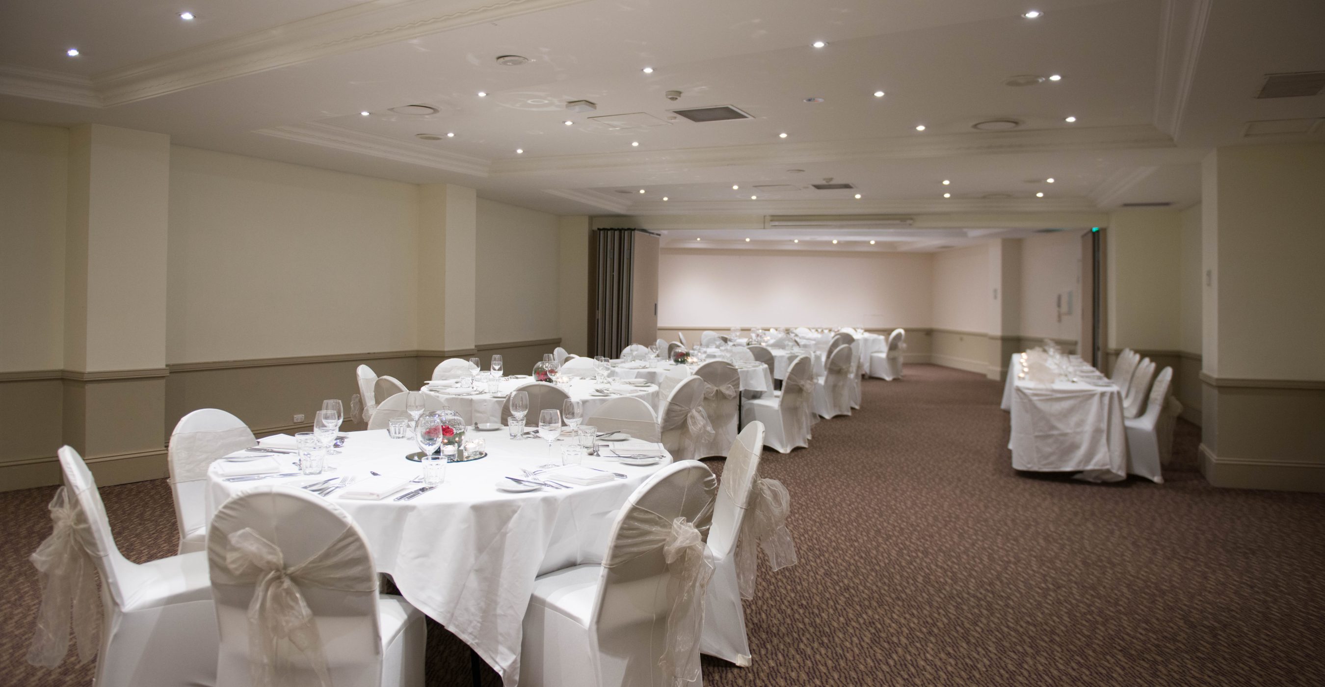 Banquet hall with round tables set with white linens and chairs, beneath a recessed ceiling with lights.
