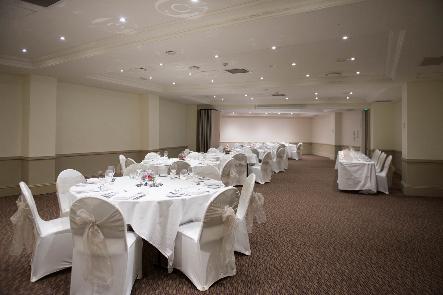 Banquet hall with round tables covered in white linens, white chair covers, and recessed ceiling lights.