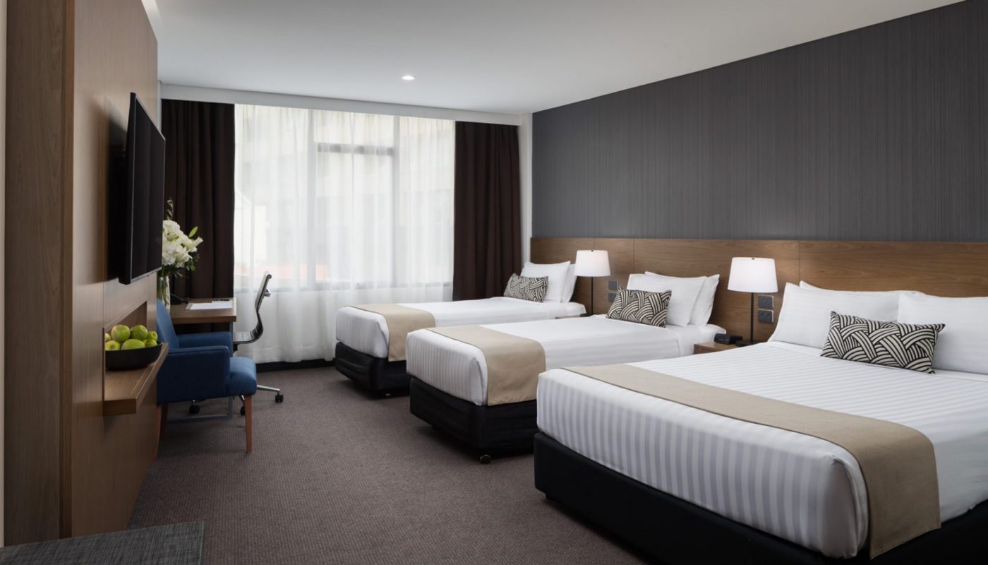 A modern hotel room with two beds, dark wood-paneled walls, a desk and chair, and large curtained windows.