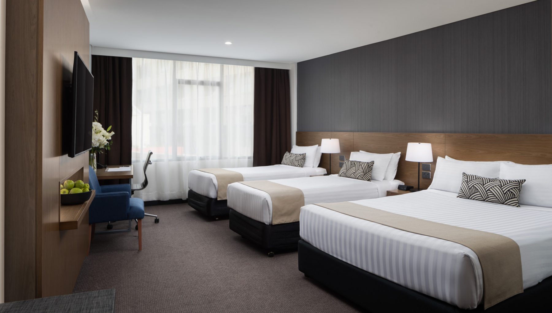 A modern hotel room with two beds, dark wood-paneled walls, a desk and chair, and large curtained windows.