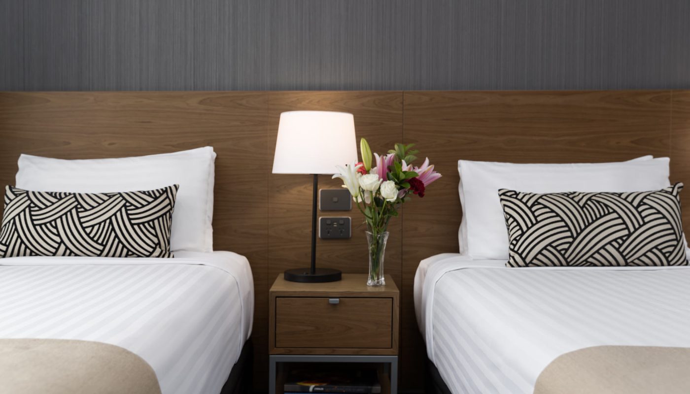 Two neatly made beds flank a nightstand with a lamp and flowers, against a wood headboard and gray wall.