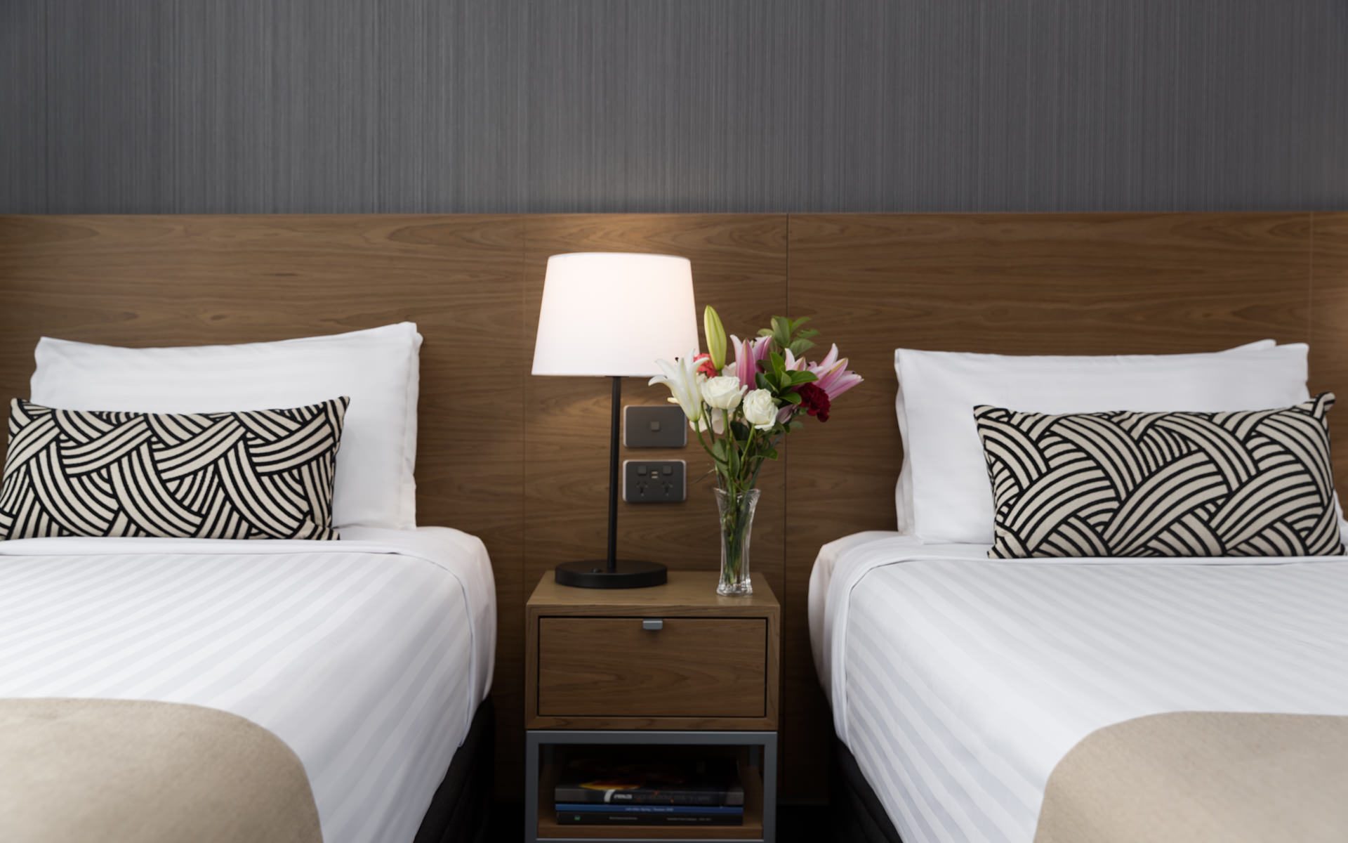 Two neatly made beds flank a nightstand with a lamp and flowers, against a wood headboard and gray wall.