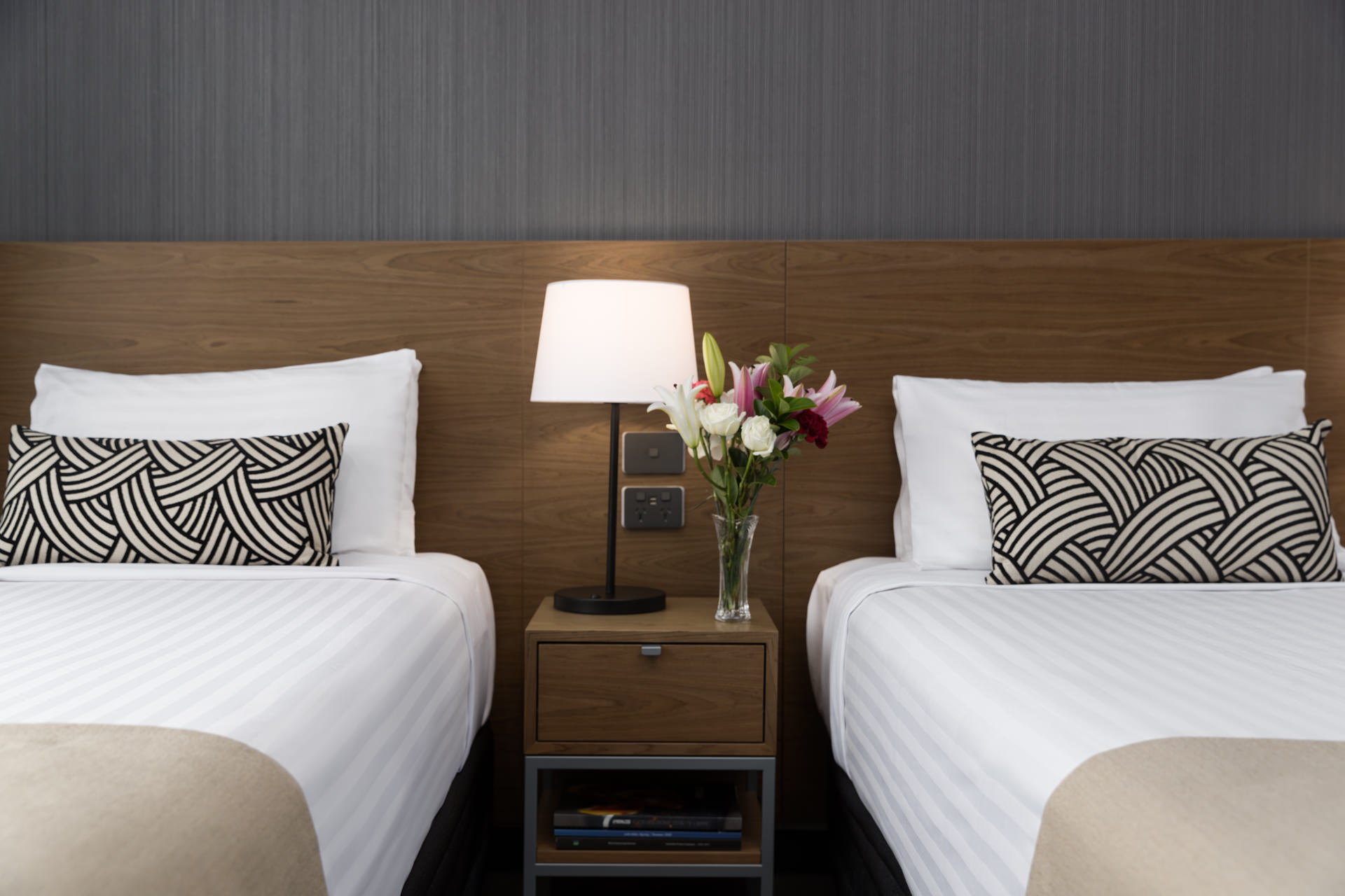 Two neatly made beds flank a nightstand with a lamp and flowers, against a wood headboard and gray wall.