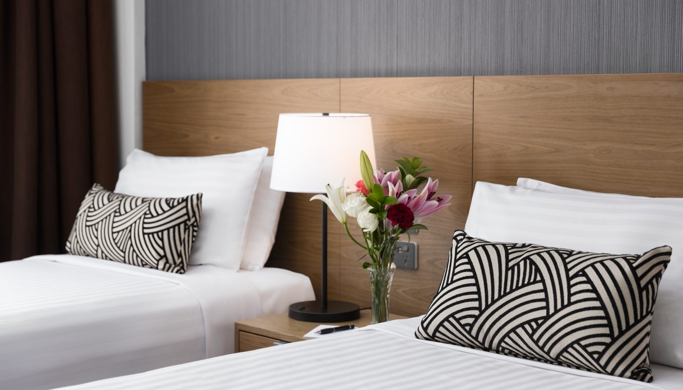 Modern bedroom with a white bed, black-and-white patterned cushions, wooden headboard, wall lamp, and flowers in a vase.