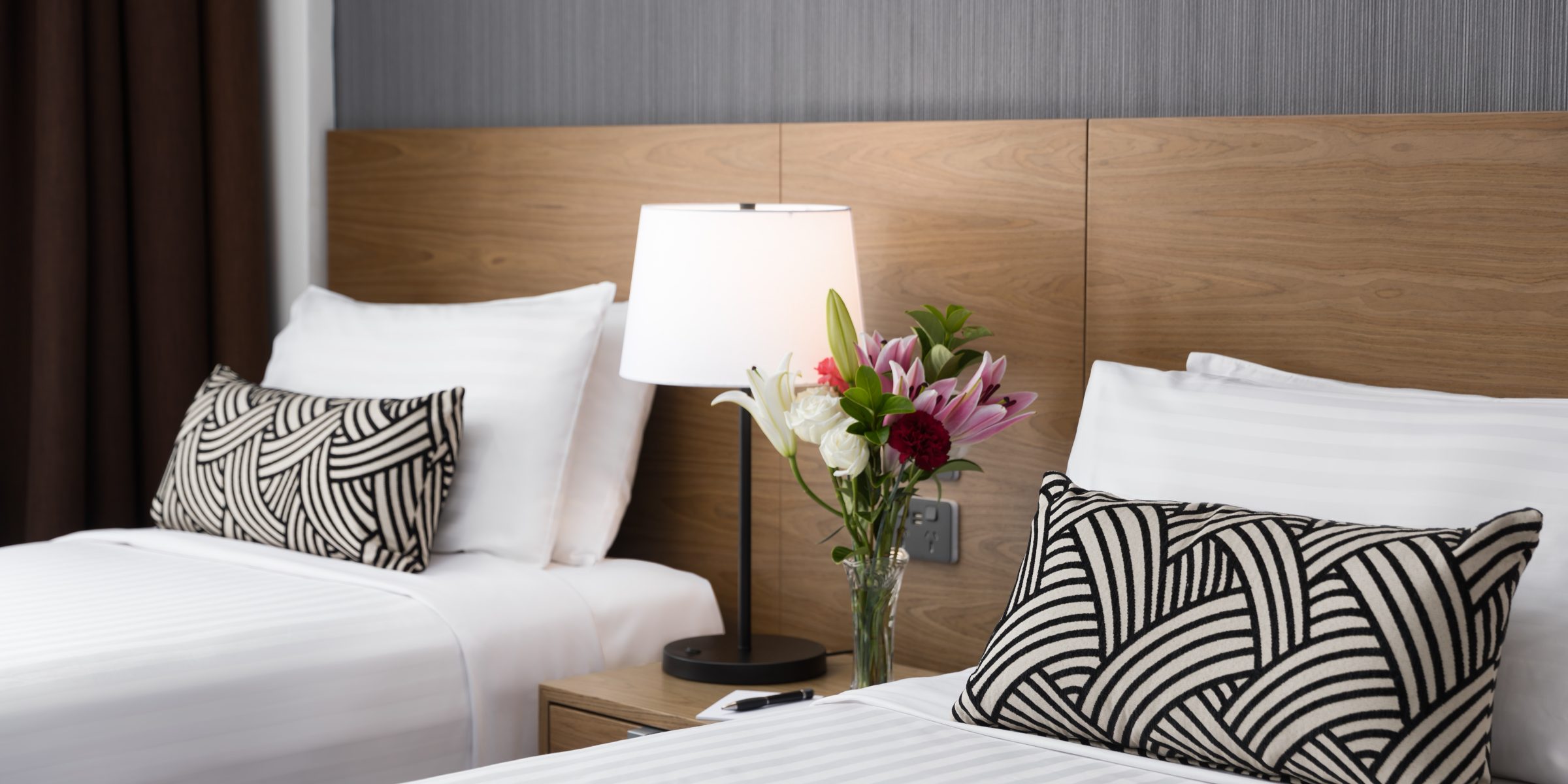 Modern bedroom with a white bed, black-and-white patterned cushions, wooden headboard, wall lamp, and flowers in a vase.