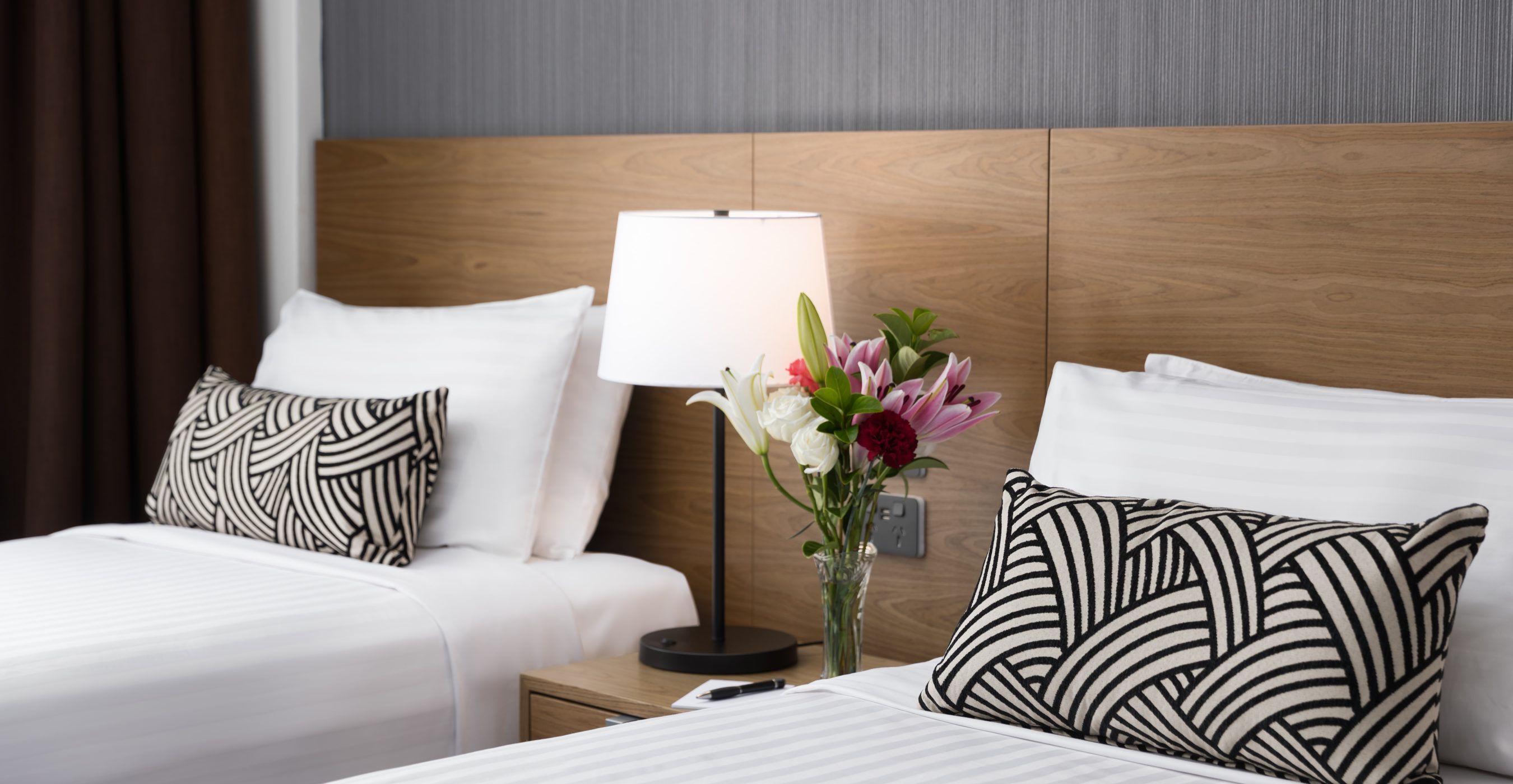Modern bedroom with a white bed, black-and-white patterned cushions, wooden headboard, wall lamp, and flowers in a vase.