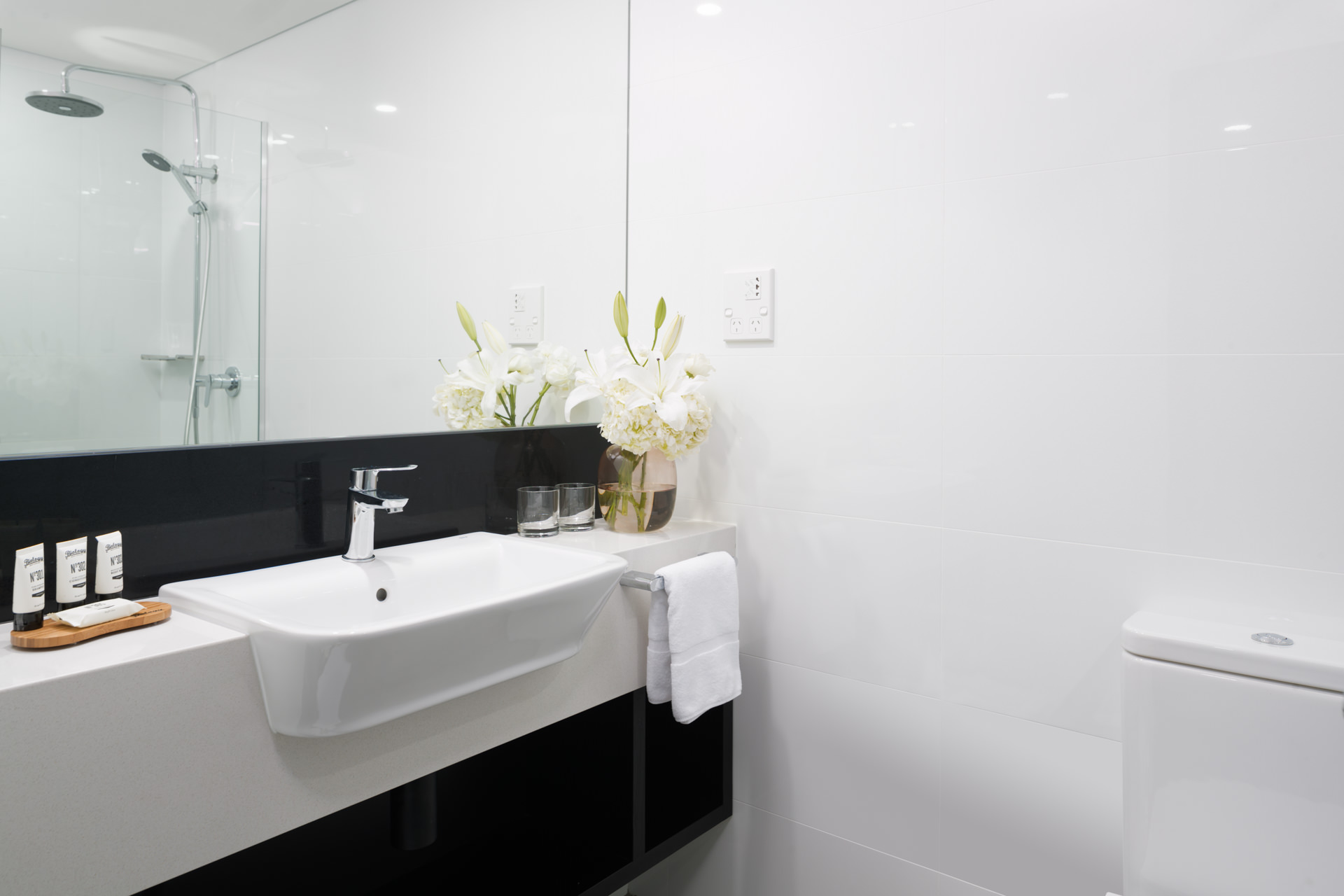 Modern bathroom with a white sink beneath a large mirror, toiletries and a plant on the counter, and a toilet beside it.