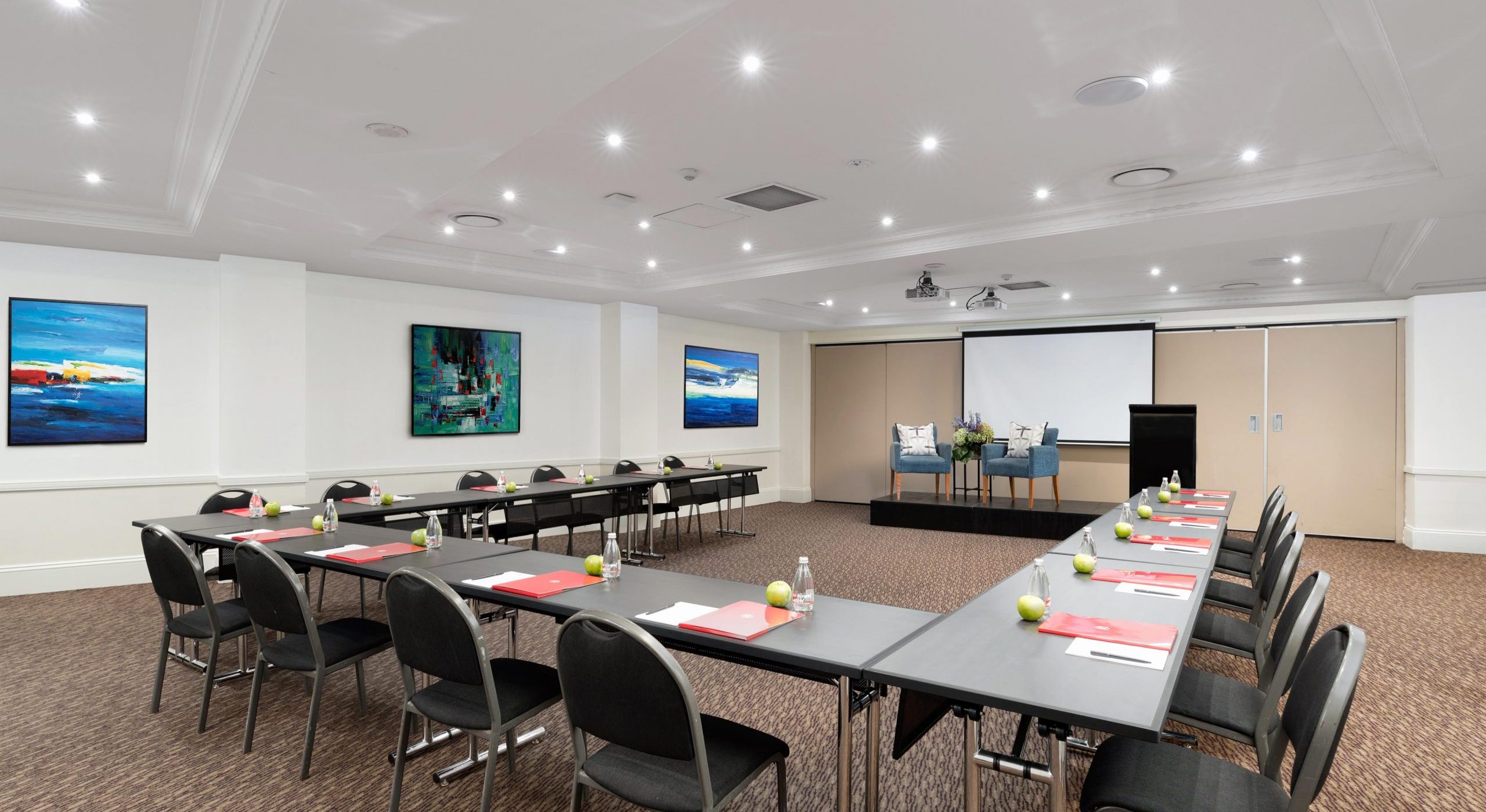 Conference room with long tables, black chairs, a projector screen, and recessed ceiling lights.