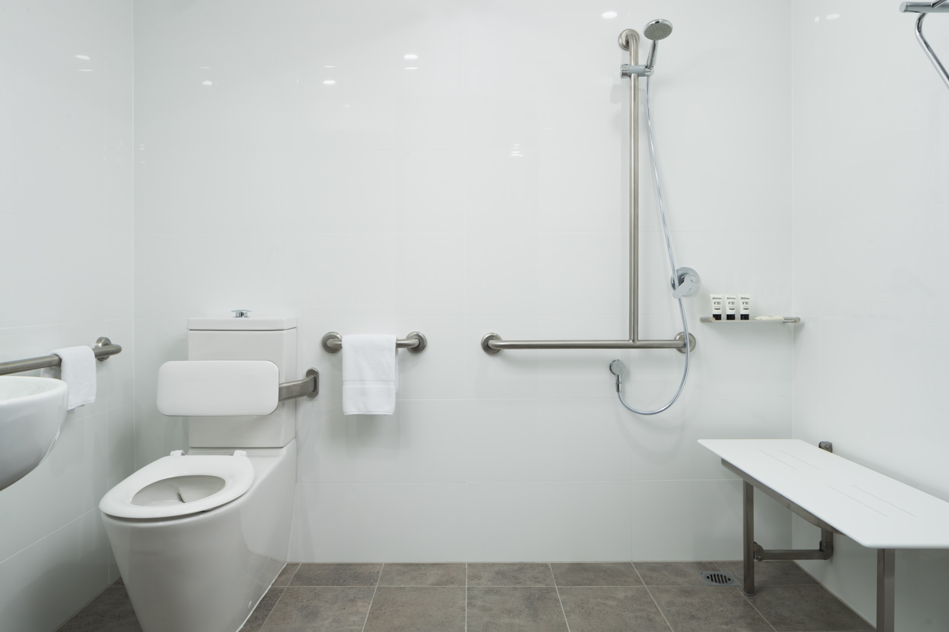 White bathroom with a toilet, wall-mounted grab bars, handheld shower, and low bench.