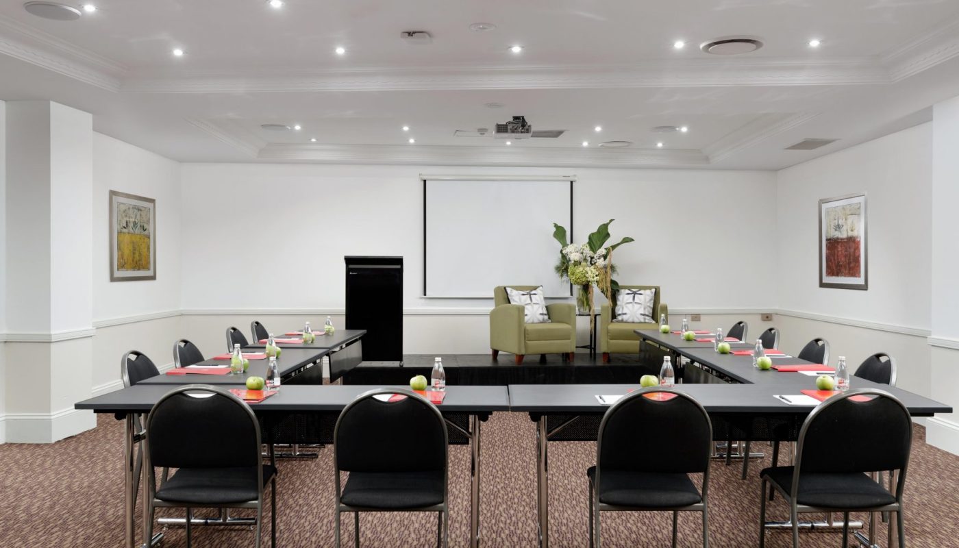 A conference room with U-shaped tables, black chairs, a projector screen, and two armchairs on a small stage.
