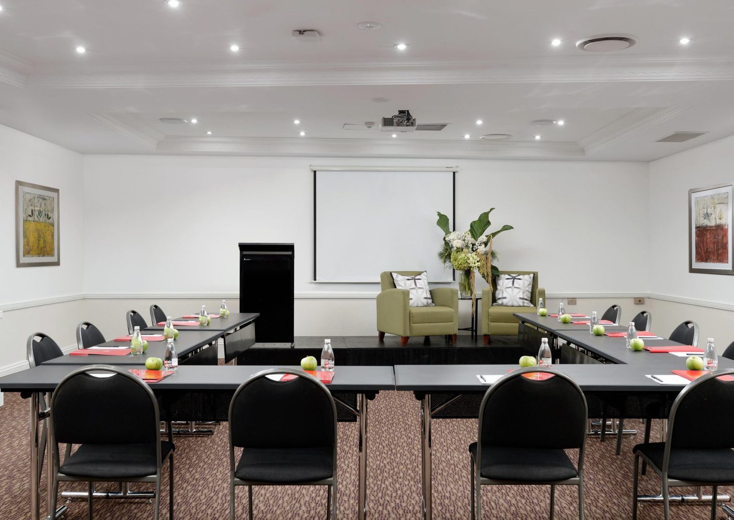 A conference room with U-shaped tables, black chairs, a projector screen, and two armchairs on a small stage.