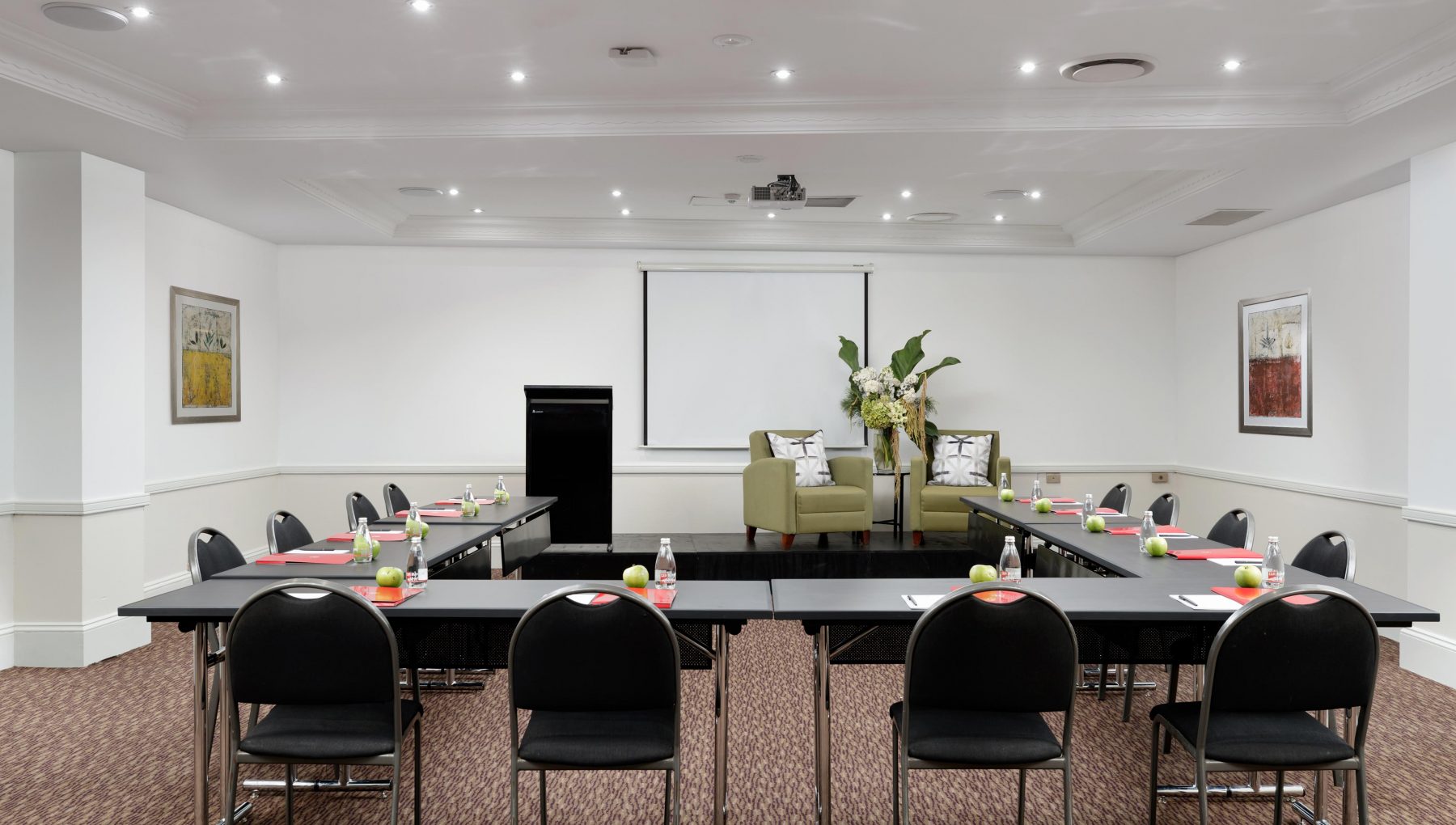 A conference room with U-shaped tables, black chairs, a projector screen, and two armchairs on a small stage.