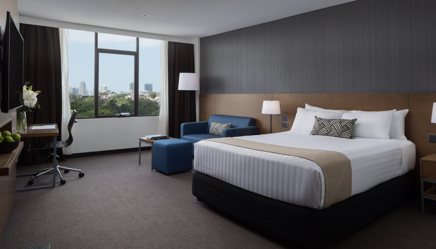 Modern hotel room with a large bed, blue sofa, desk, and floor-to-ceiling window overlooking the city.