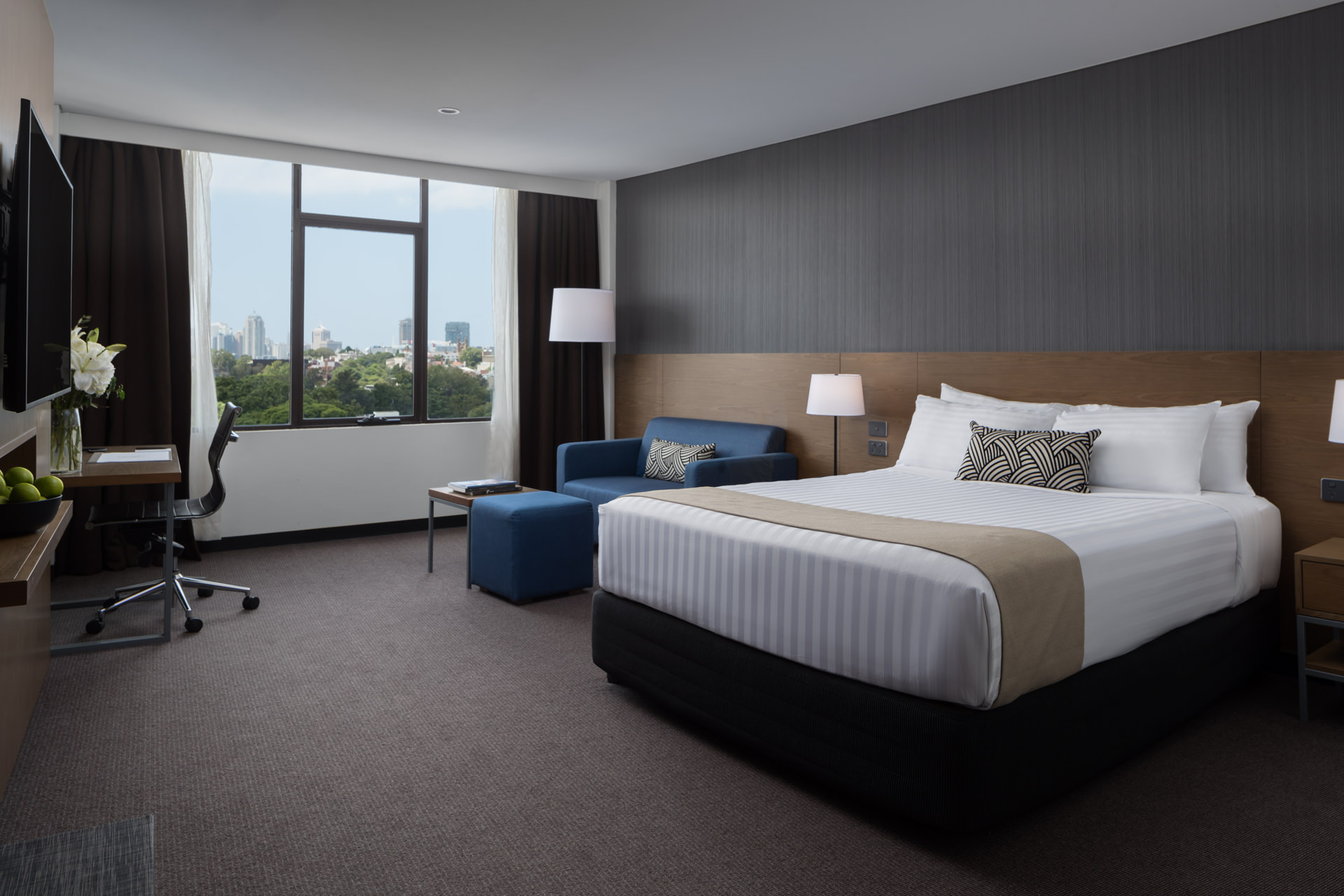 Modern hotel room with a large bed, blue sofa, desk, and floor-to-ceiling window overlooking the city.