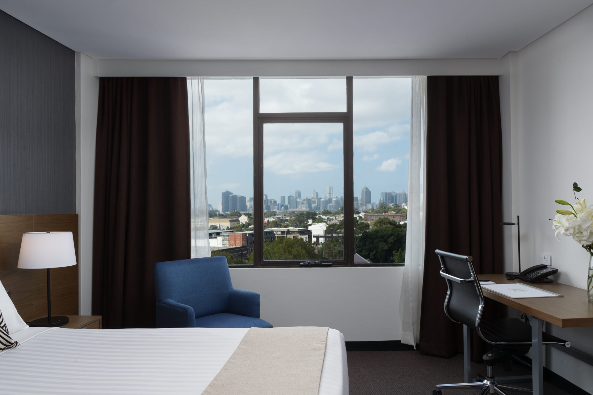 Bedroom with a bed, blue armchair, and desk beside a large window overlooking a city skyline.