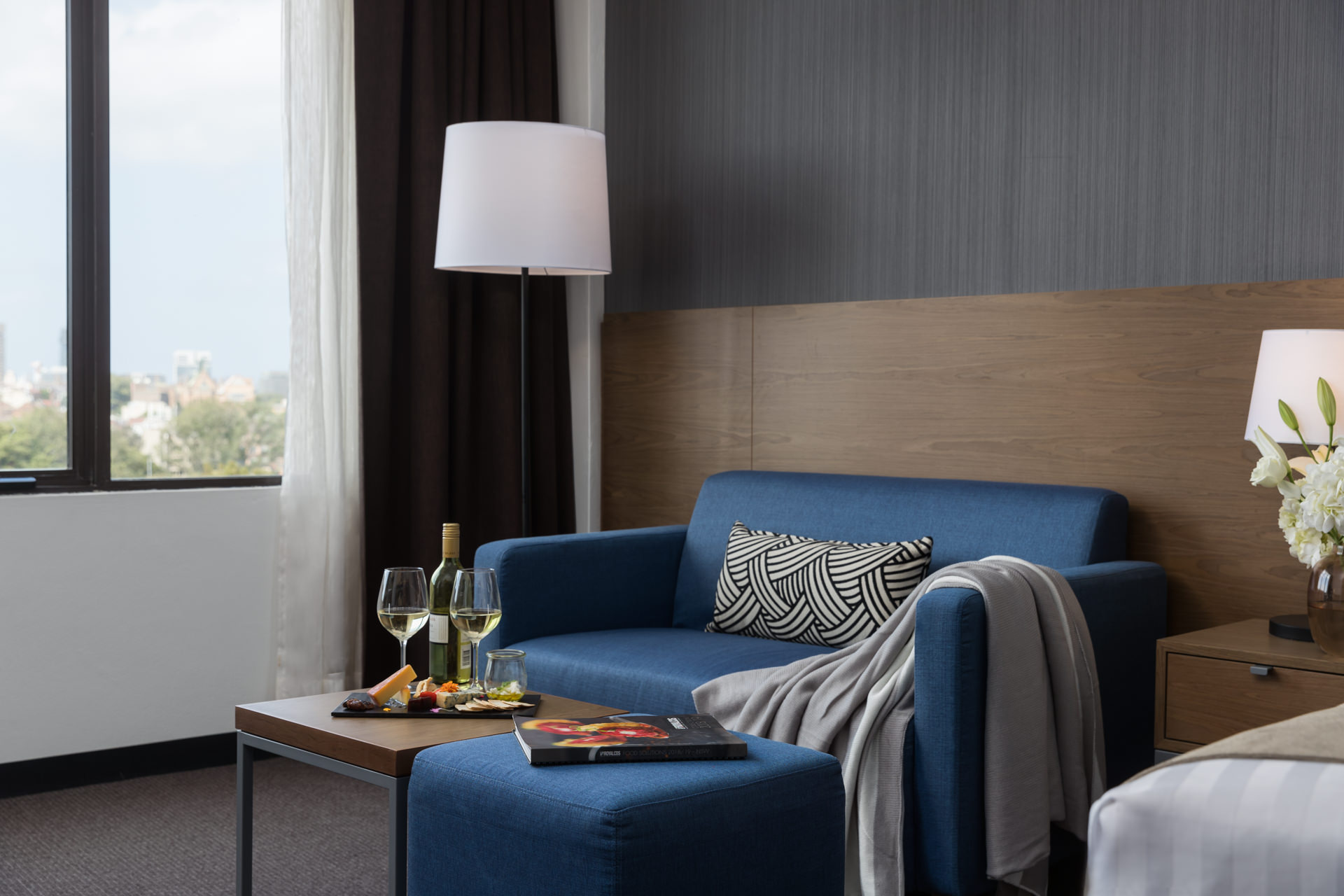 Blue loveseat with patterned cushions and a gray throw, matching ottoman, floor lamp, and large window.