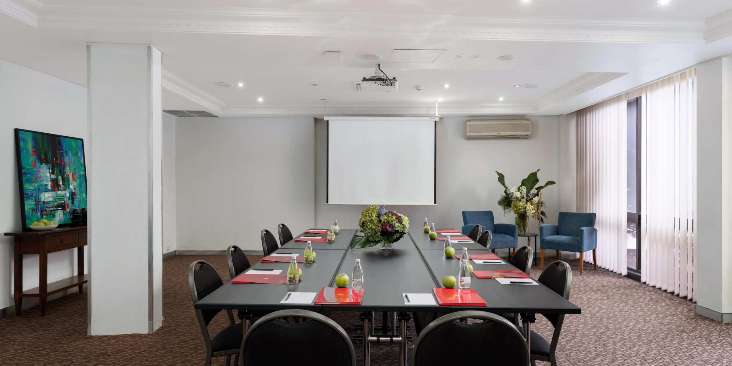 A conference room with a long table and chairs, a projection screen, plants, and tall windows.