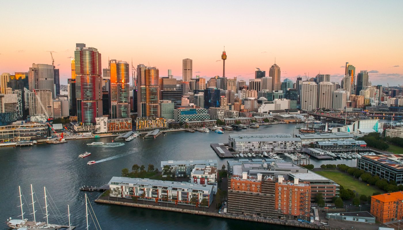 City skyline at sunset overlooking a harbor with boats, piers, and waterfront buildings.