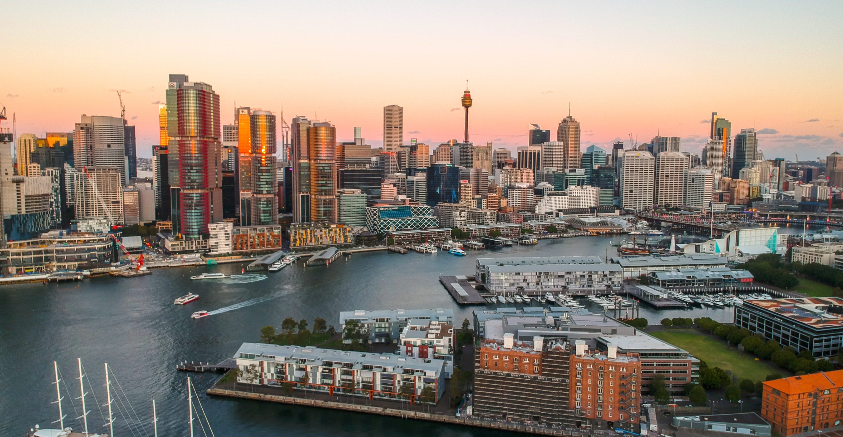 City skyline at sunset overlooking a harbor with boats, piers, and waterfront buildings.