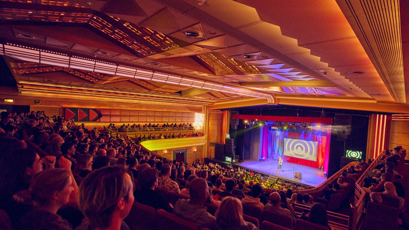 Packed auditorium viewed from above, with a seated audience facing a brightly lit stage beneath an ornate ceiling.