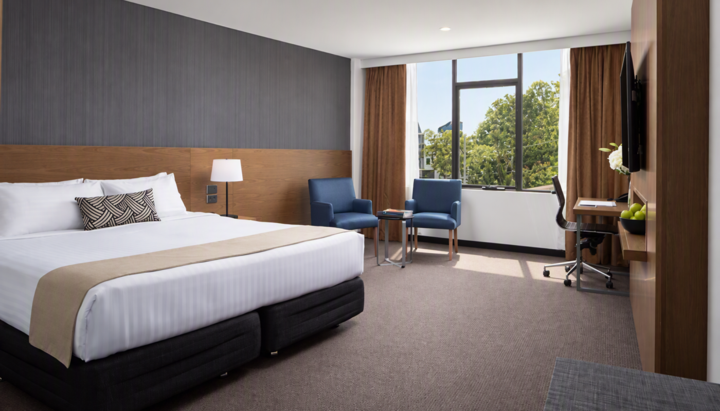 Modern hotel room with a large white bed, two blue chairs by a sunlit window, and a desk with a wall-mounted TV.