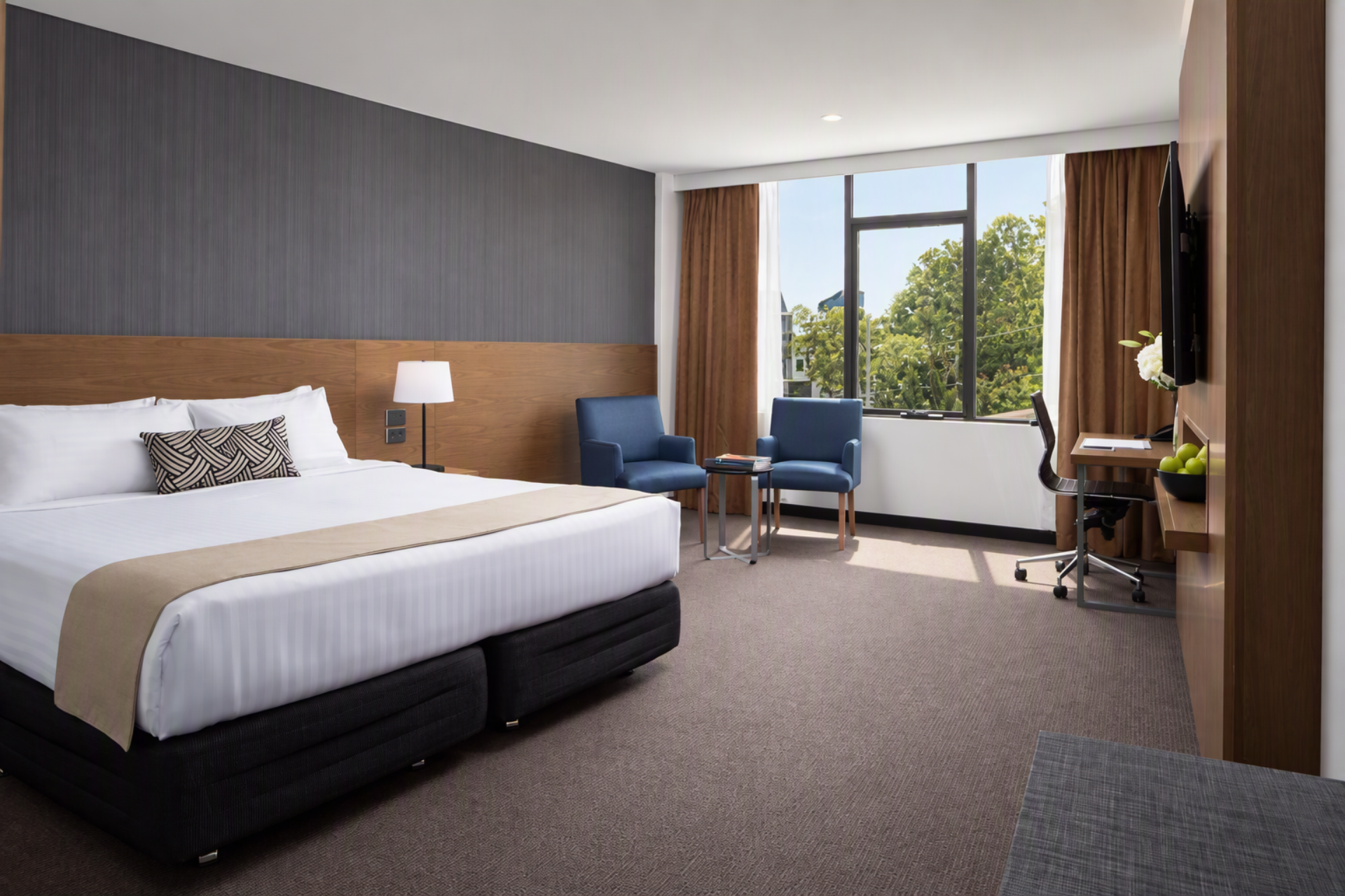 Modern hotel room with a large white bed, two blue chairs by a sunlit window, and a desk with a wall-mounted TV.