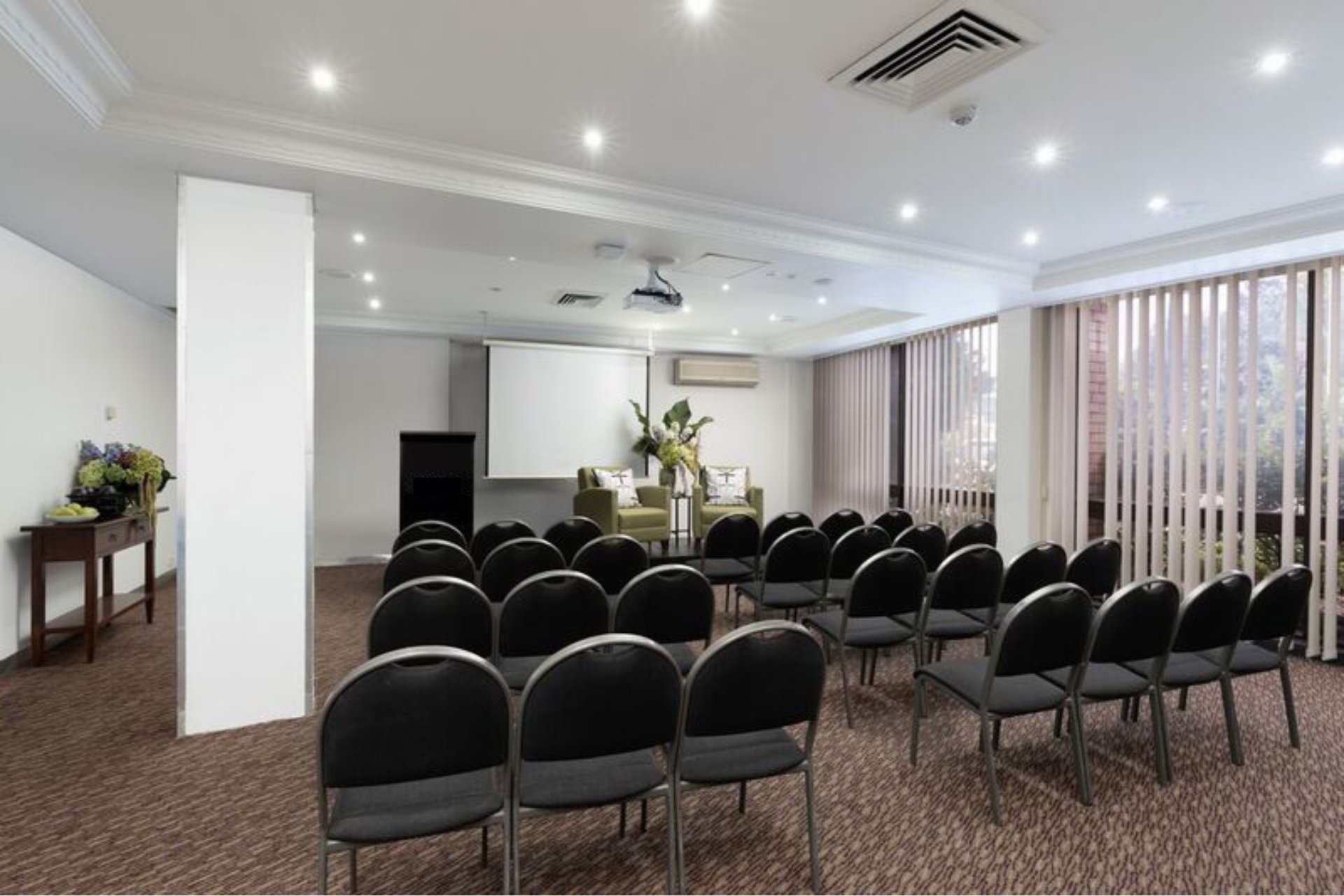 Rows of black chairs face a projector screen and lectern in a bright meeting room with vertical blinds.