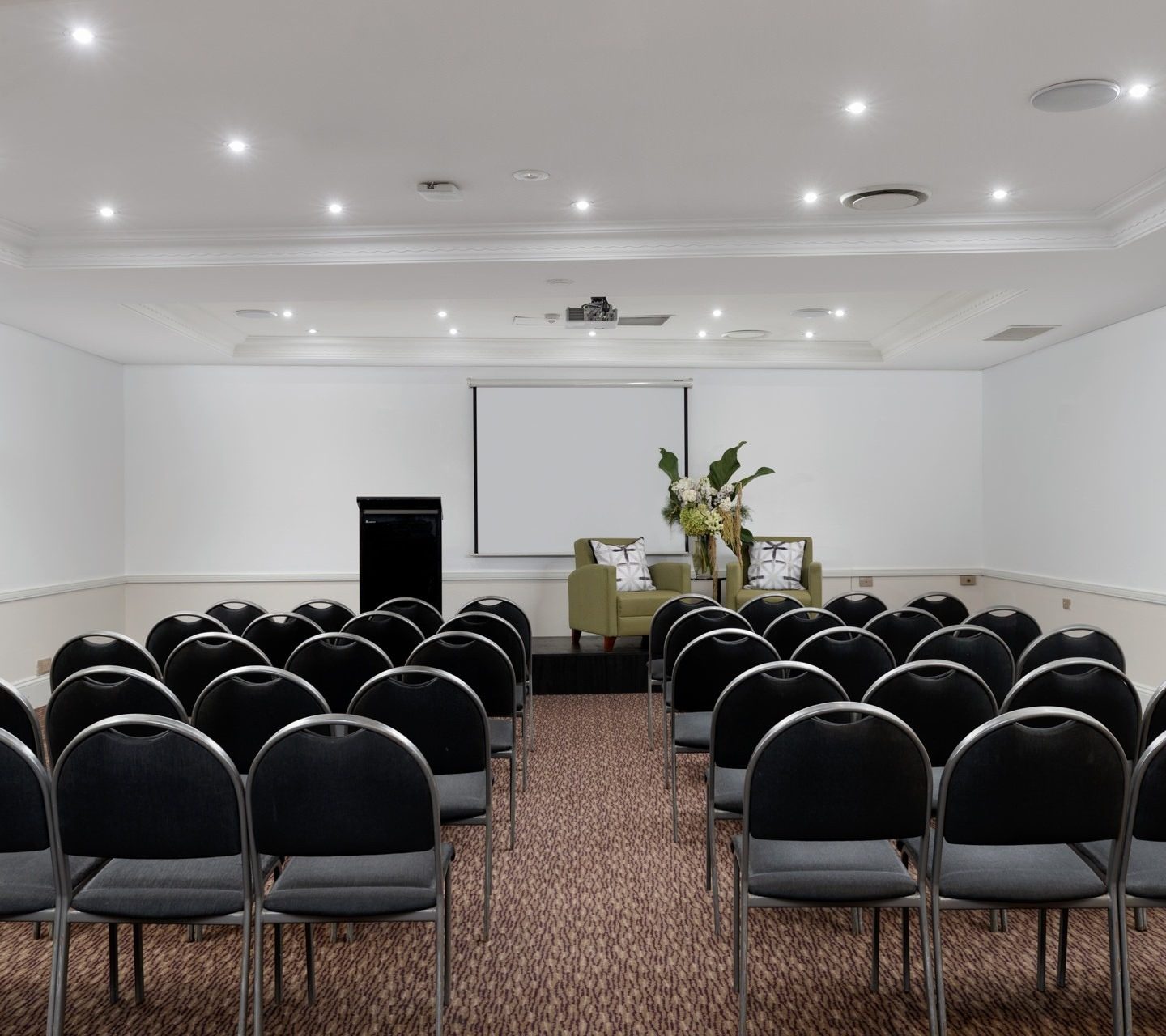 Rows of black chairs face a small stage with two green armchairs, a flower arrangement, projector screen, and lectern.
