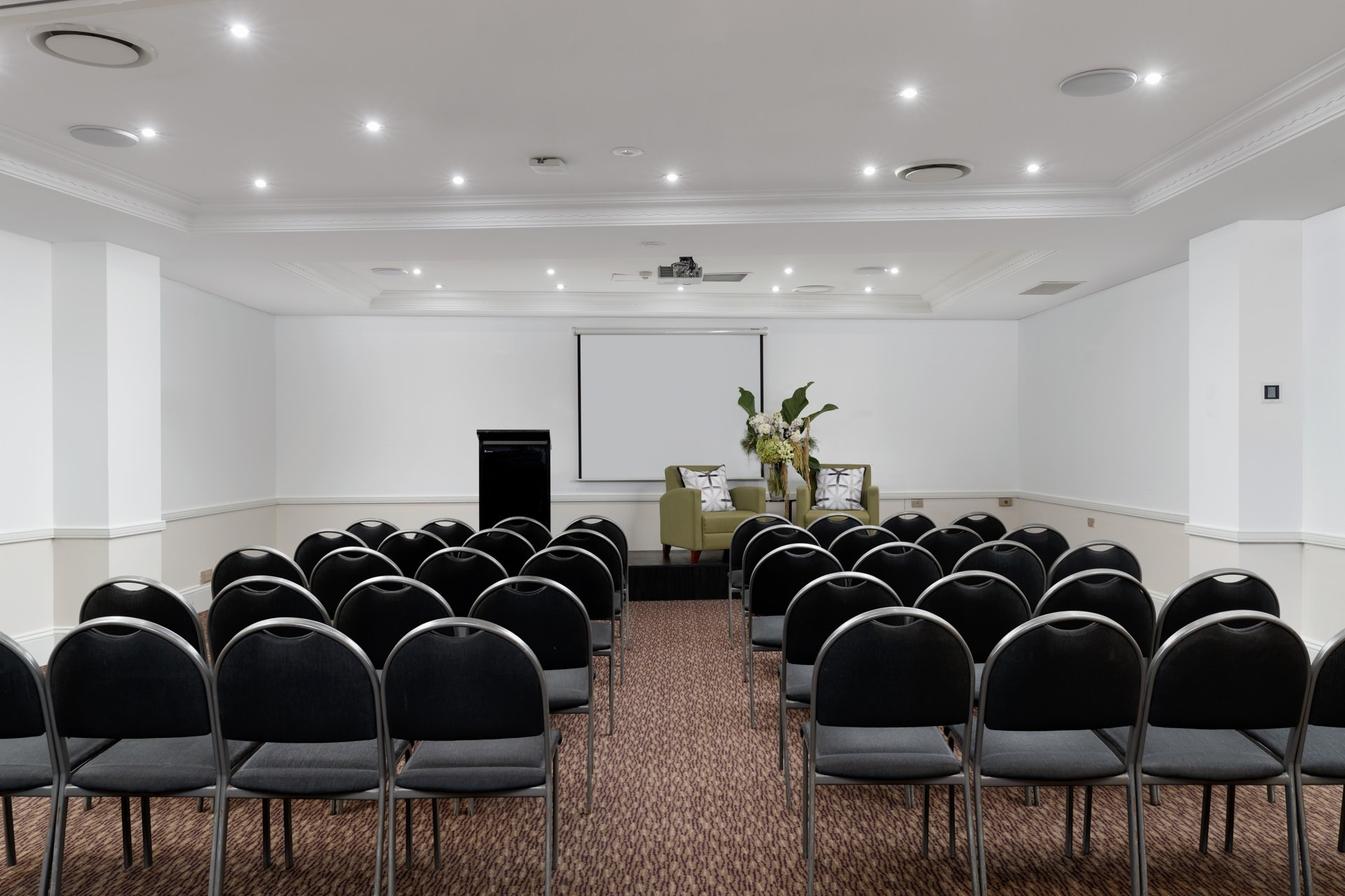 Rows of black chairs face a small stage with two green armchairs, a flower arrangement, projector screen, and lectern.