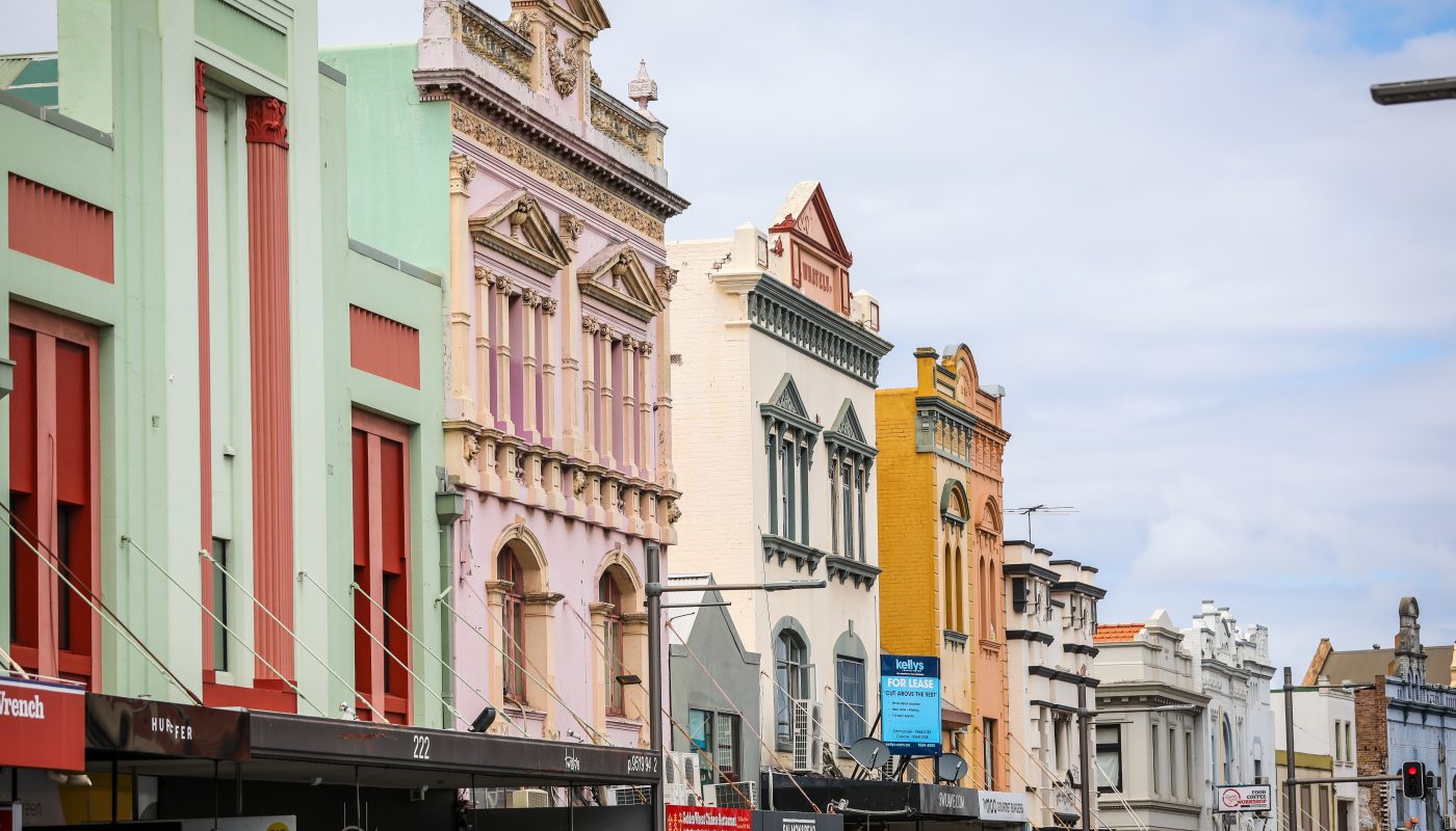 A row of colorful, ornate shopfronts lines a city street beneath a cloudy sky.