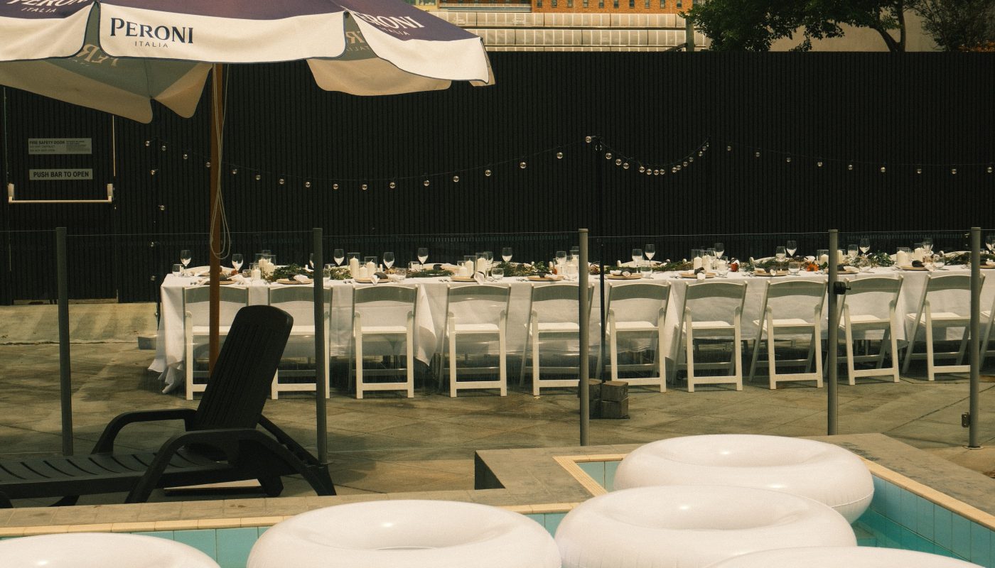 A poolside dining area is set with long tables, white chairs, string lights, large umbrellas, and floating loungers.