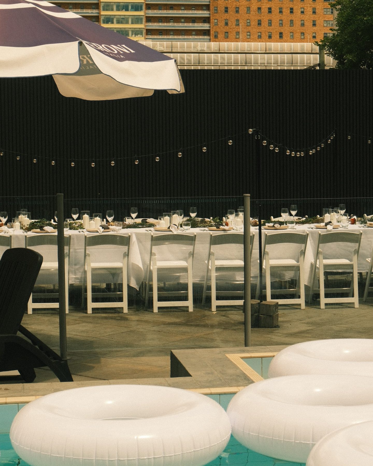 A poolside dining area is set with long tables, white chairs, string lights, large umbrellas, and floating loungers.