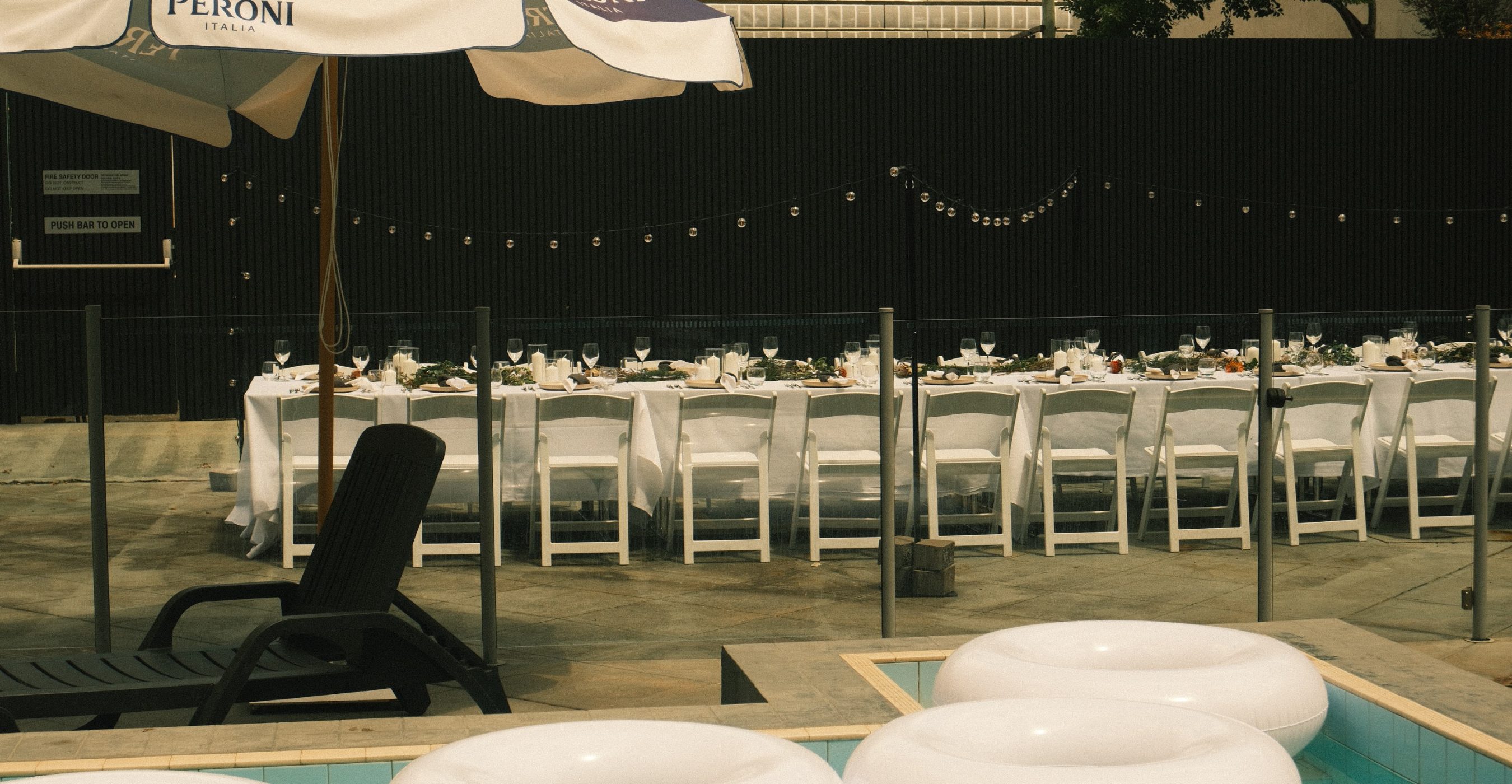 A poolside dining area is set with long tables, white chairs, string lights, large umbrellas, and floating loungers.