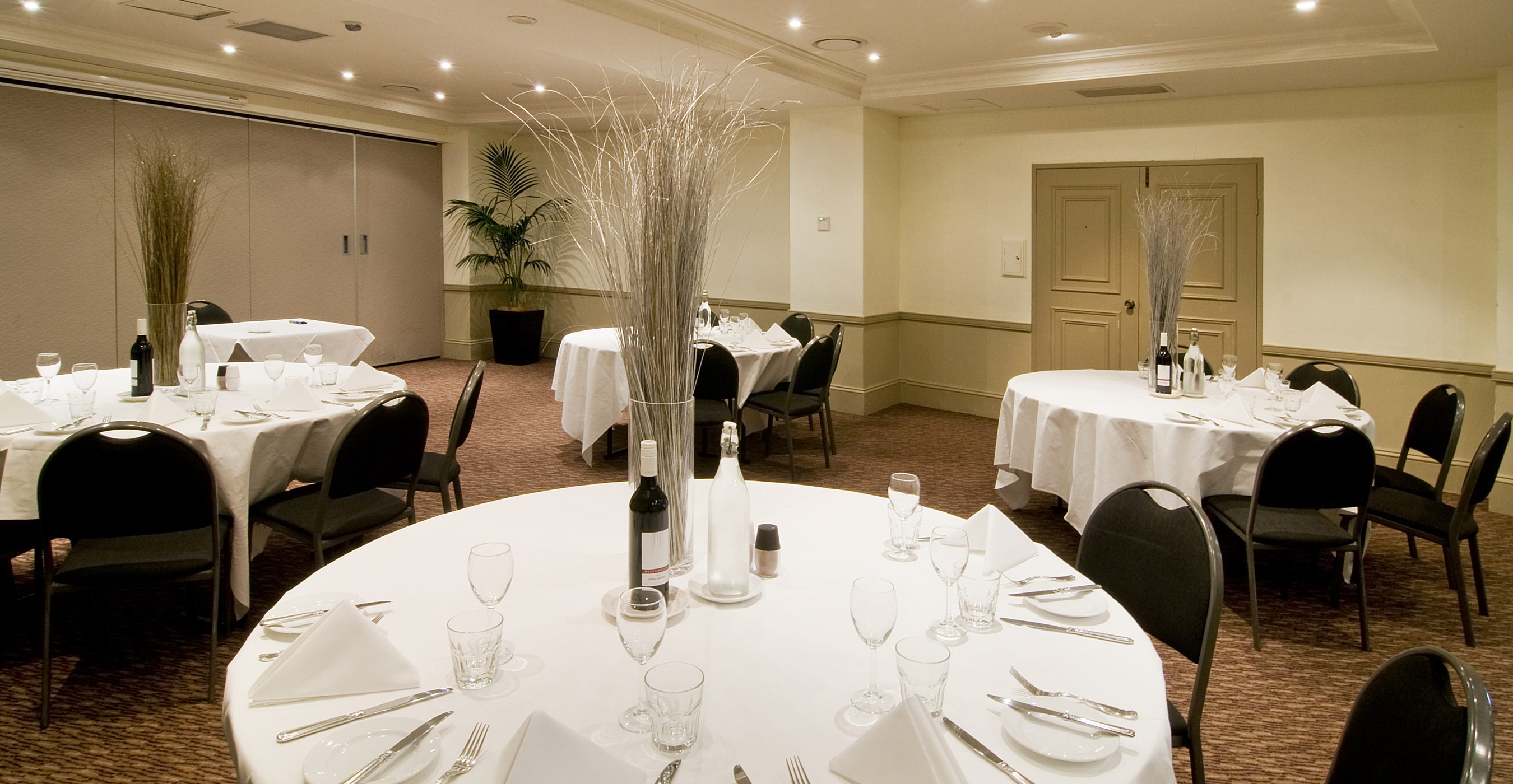 A banquet room with round tables set with white linens, glassware, candles, and dark chairs.