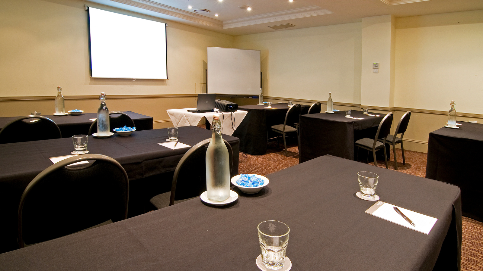 Conference room with black tablecloths, chairs, water glasses and bottles, blue plates, and a projection screen.