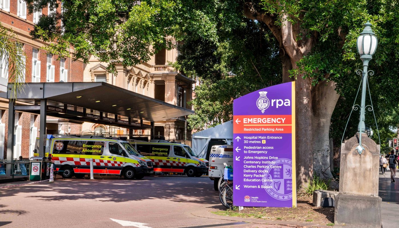Ambulances parked beside a purple emergency sign outside a brick building, partly shaded by a large tree.