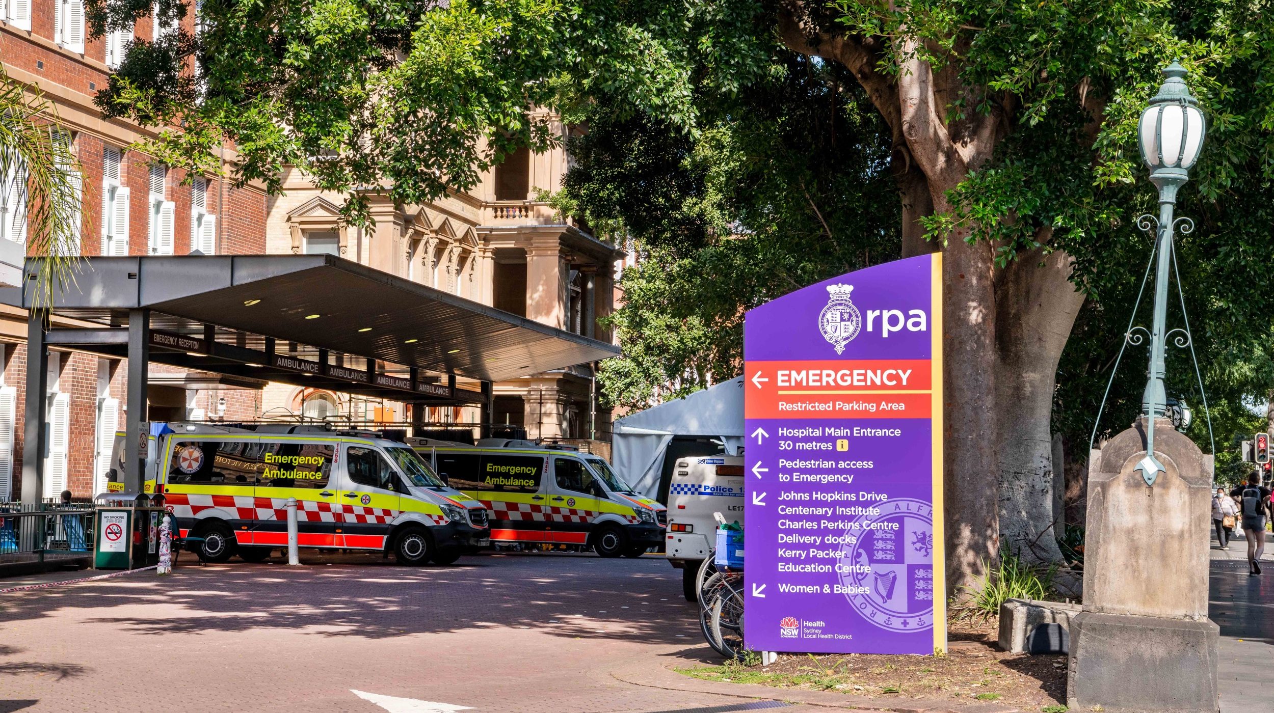 Ambulances parked beside a purple emergency sign outside a brick building, partly shaded by a large tree.