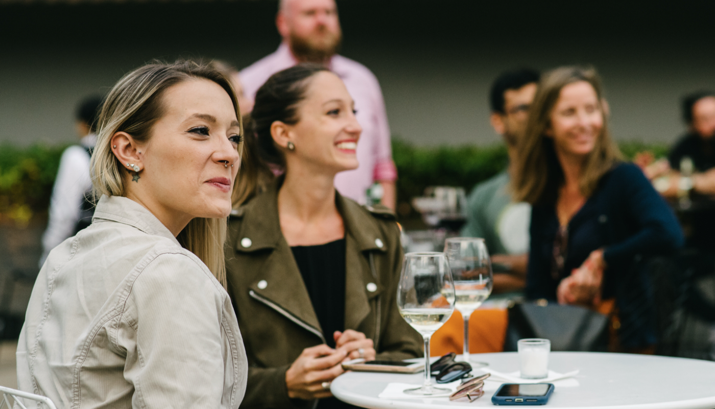 Two women sit at a white outdoor café table with drinks as other guests mingle behind them.