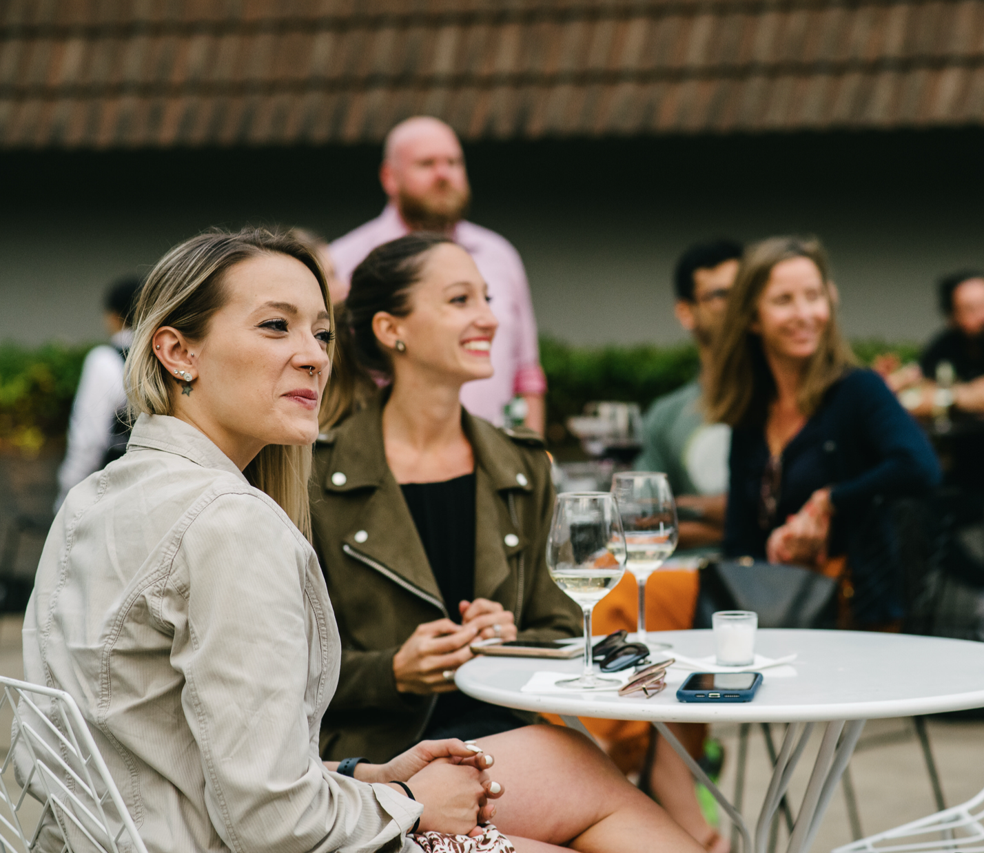 Two women sit at a white outdoor café table with drinks as other guests mingle behind them.