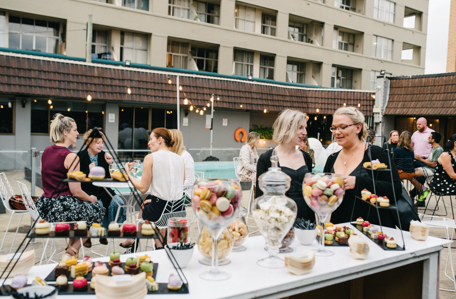 Guests mingle around a dessert table in a courtyard, with two women chatting beside bowls of sweets.