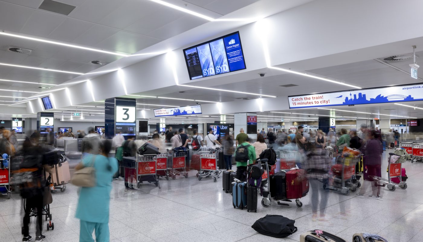 Travelers queue at airport check-in counters with suitcases beneath overhead flight information screens.