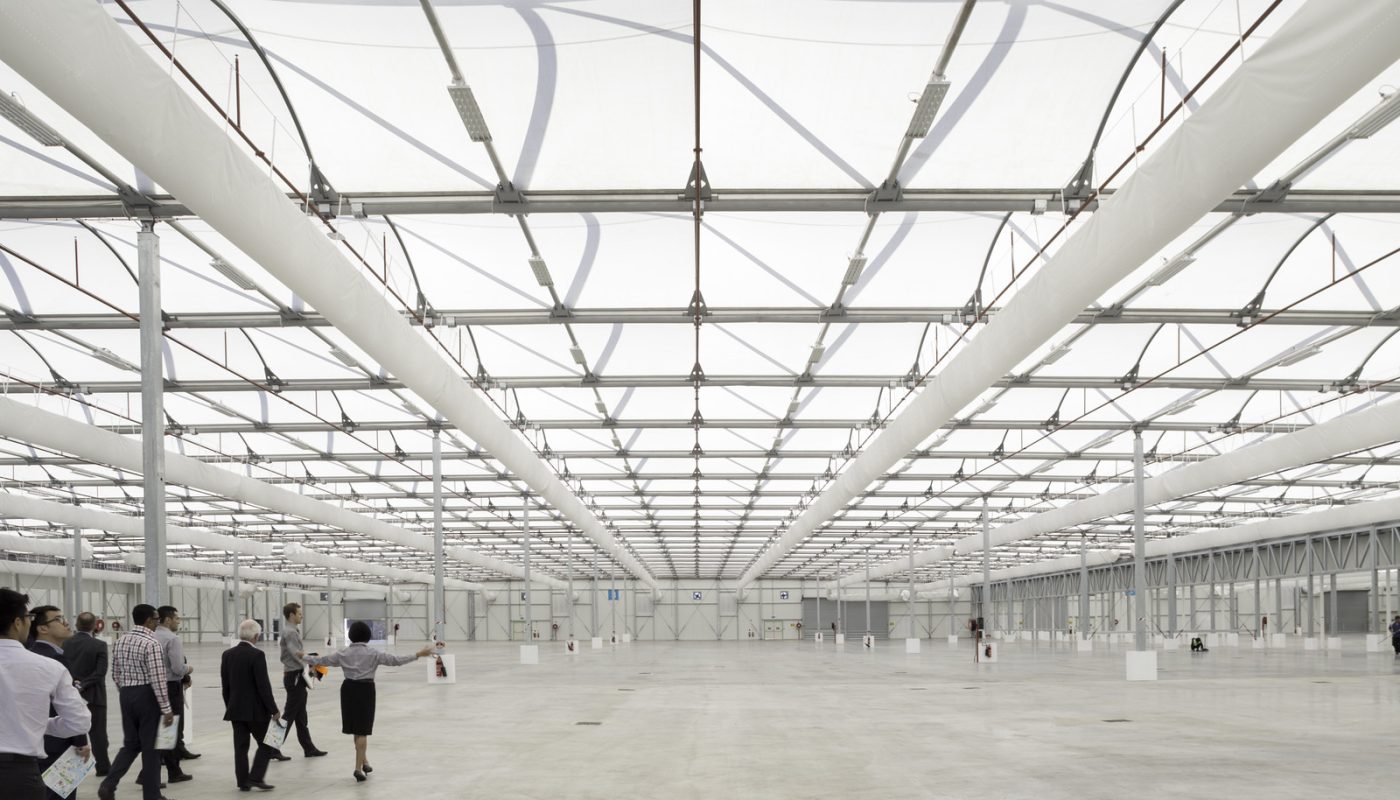 A vast, bright industrial hall with a grid of overhead beams and a few people walking across the open floor.