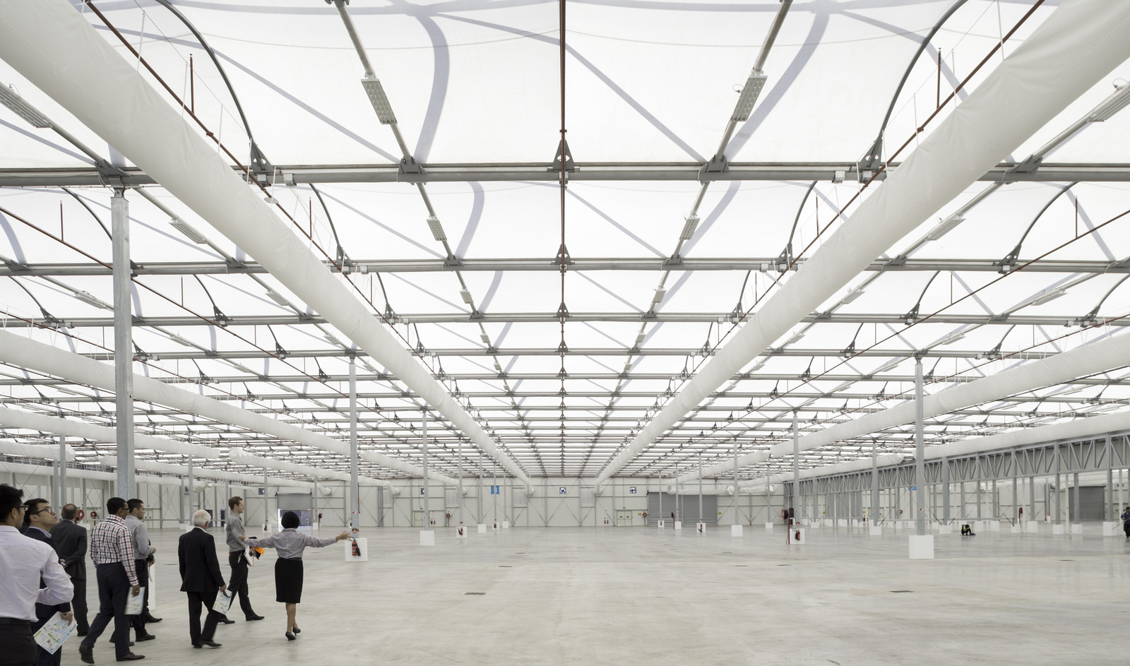 A vast, bright industrial hall with a grid of overhead beams and a few people walking across the open floor.