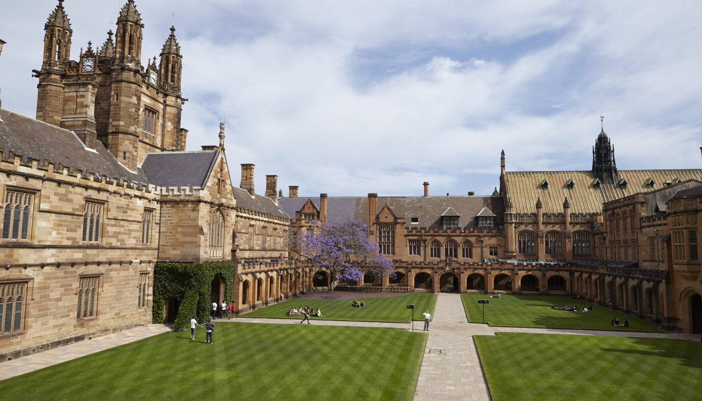 Stone college courtyard with a central path across green lawns, surrounded by historic buildings and towers under a cloudy sky.