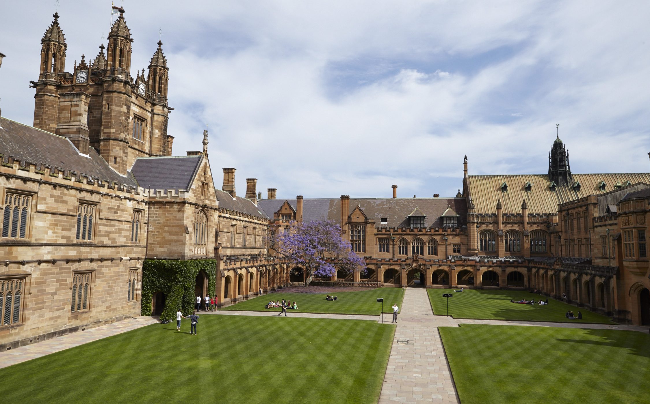 Stone college courtyard with a central path across green lawns, surrounded by historic buildings and towers under a cloudy sky.
