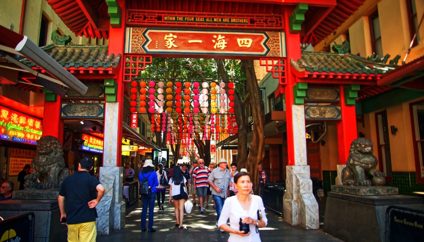 Crowds walk beneath an ornate red-and-green temple gate hung with colorful lanterns, with a woman in the foreground.