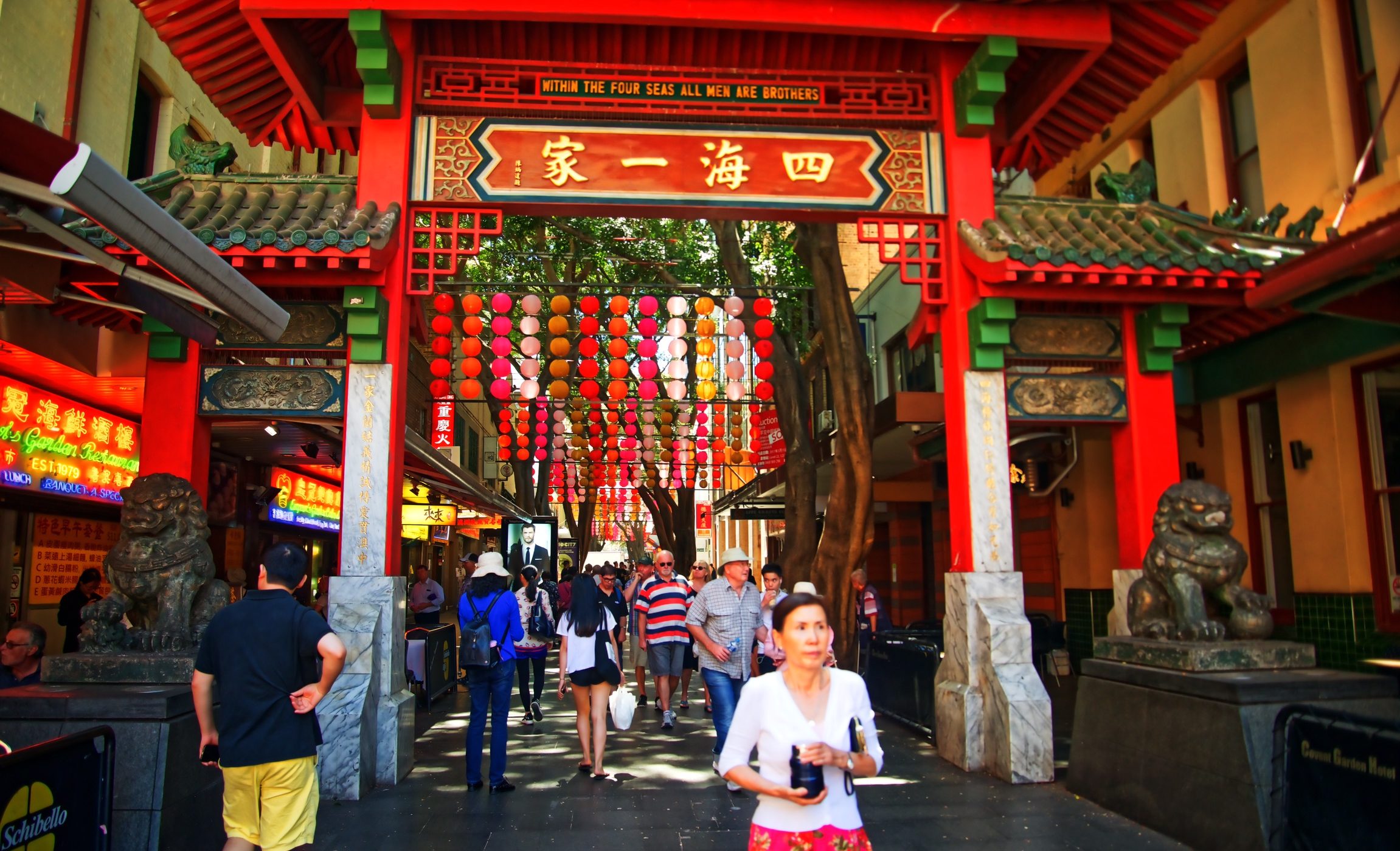 Crowds walk beneath an ornate red-and-green temple gate hung with colorful lanterns, with a woman in the foreground.