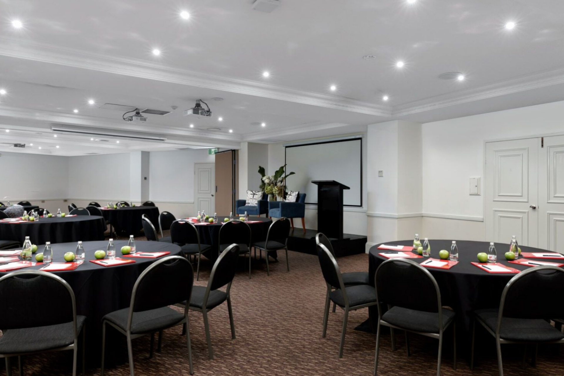 A conference room with round tables set with dishes, black chairs, and a podium beside a projection screen.