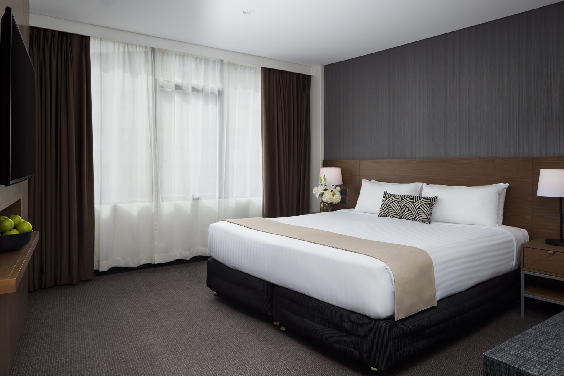 A modern bedroom with a king-size bed, white linens, beige runner, dark gray wall, and floor-to-ceiling curtains.