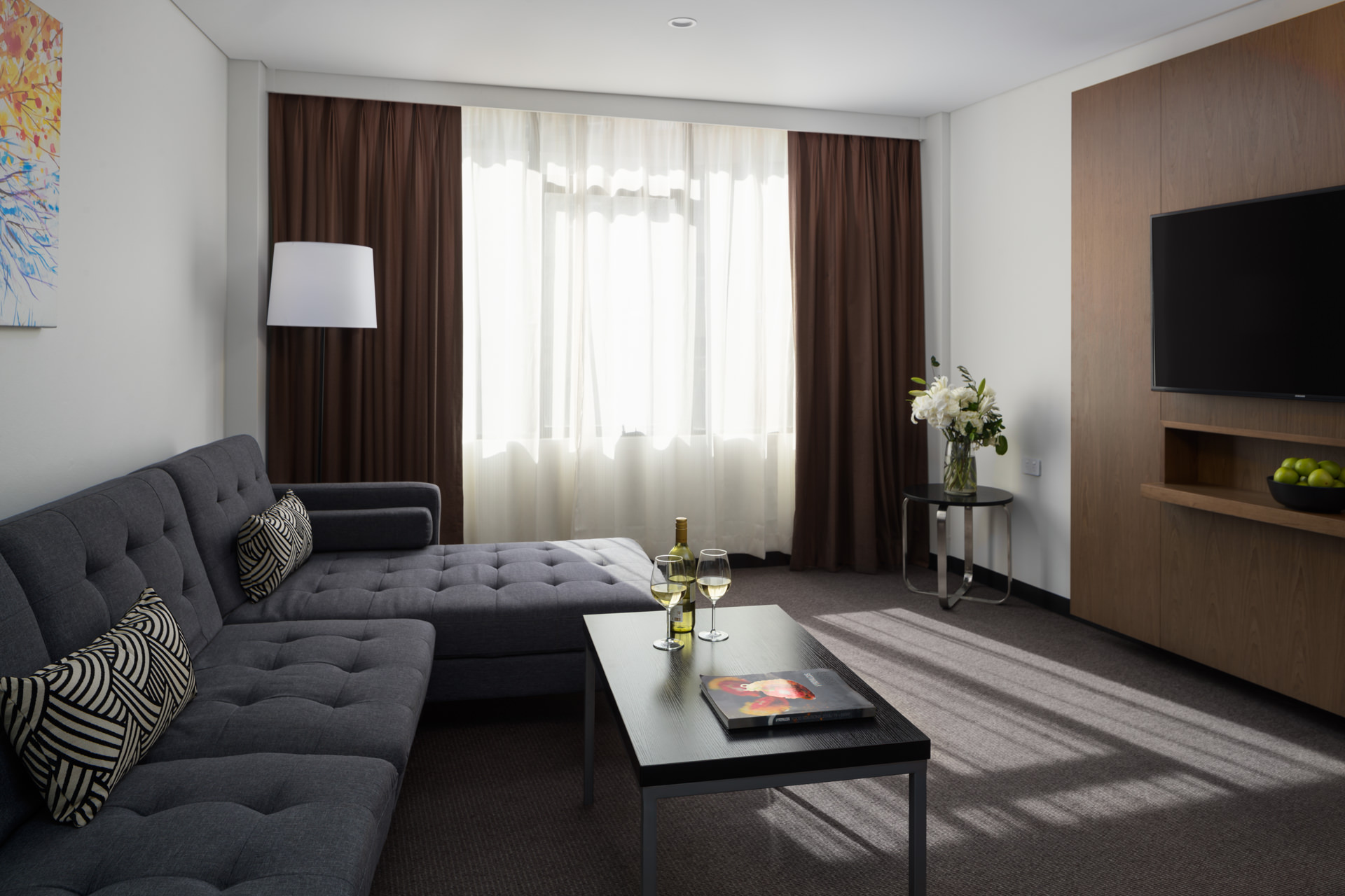 Modern living room with a gray sectional sofa, black coffee table, floor lamp, sheer curtains, and wall-mounted TV.